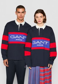 Marine langermet polo med røde og blå striper, med hvit krage og logoer. Båret av to modeller i uformell attire.
