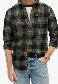 Camicia a maniche lunghe a quadri in navy, verde e crema; miscela di cotone. Presenta una tasca sul petto e un discreto logo ricamato. Indossata sopra una t-shirt bianca.