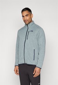 Casaco polar em azul claro com colarinho alto e fecho de correr, apresentando bolsos laterais e detalhes em azul-marinho, feito de tecido de textura suave.