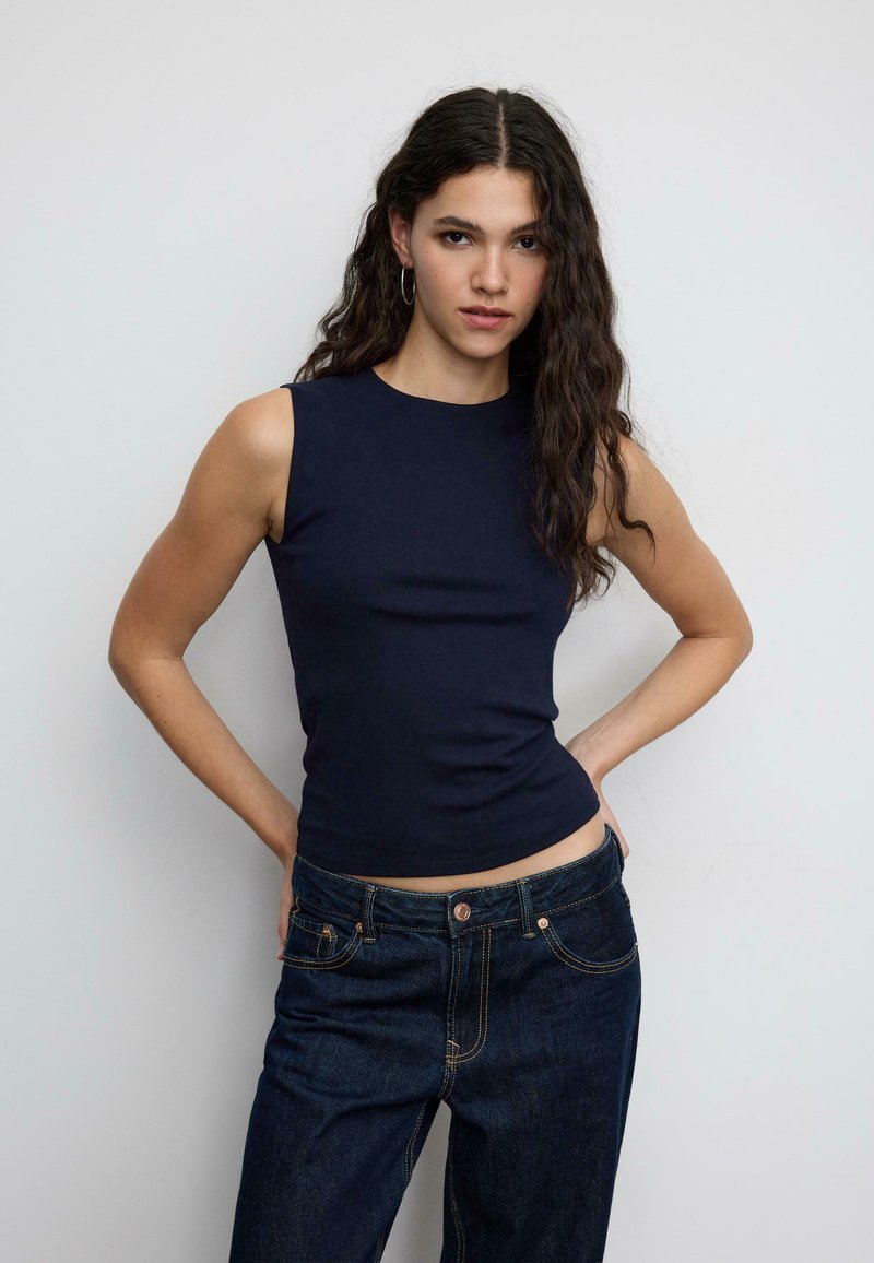 Marineblaues ärmelloses Oberteil aus einem glatten Stoff, mit rundem Ausschnitt und figurbetonter Passform, kombiniert mit dunklen Jeans.