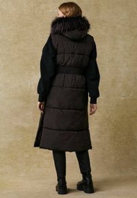 Langer, dunkler Puffermantel mit Kapuze aus Fell, strukturierten schwarzen Ärmeln, elastischem Taillenbund und seitlichen Schlitzen, getragen über schwarzen Hosen und Stiefeln.