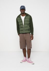 Roheline puhvvest ribidega varrukatega, valge T-särk, pruunid cargo-lühikesed püksid ja roosad kõrged tennisekingad. Imitab muretud kihilist riietust.