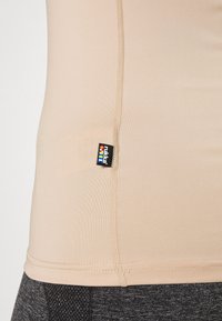 Beige idrottstopp i stretchig tyg med platta sömmar och en liten etikett på sidan, kombinerad med mörkgrå strukturerade leggings.