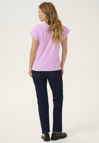 Gai violets, īsām piedurknēm T-krekls apvienots ar tumši ziliem taisnajiem džinsiem un melnām apaviem ar masīlu zoli, kurā ir teksturēta auduma virsma.