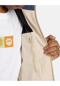 Beige jacket interior with a mesh lining, features a small zippered pocket. Person wearing a white T-shirt with colorful graphic print.