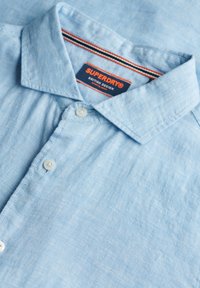 Camicia azzurro chiaro con bottoni realizzata in tessuto testurizzato. Presenta un colletto classico, pieghe sottili e un'etichetta con accenti rossi e blu.