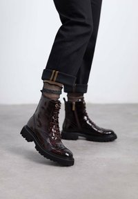 Botines de charol burdeos con un acabado texturizado, diseño con cordones y suelas de goma negra robustas. Pantalones negros ajustados con puños y calcetines de diseño visibles.