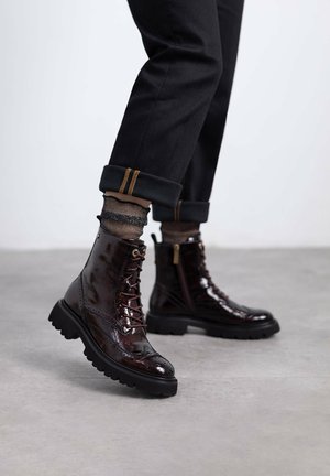 Botines de charol burdeos con un acabado texturizado, diseño con cordones y suelas de goma negra robustas. Pantalones negros ajustados con puños y calcetines de diseño visibles.