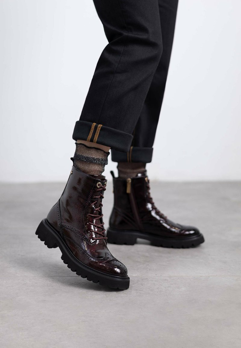 Botines de charol burdeos con un acabado texturizado, diseño con cordones y suelas de goma negra robustas. Pantalones negros ajustados con puños y calcetines de diseño visibles.