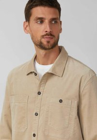 Bežová kordová košeľa s golierom, dvoma náprsníkmi a tmavými gombíkmi. Má textúrovanú látku a uvoľnený strih.
