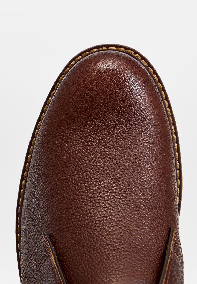 Brązowy skórzany but o teksturowanej powierzchni, zaokrąglonym czubku, kontrastowym żółtym szwie wzdłuż krawędzi i wzmocnionej konstrukcji po bokach.