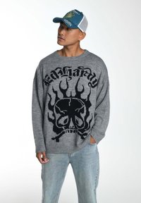 Grauer Pullover mit schwarzem Flammen- und Totenkopf-Muster, runder Ausschnitt und gerippten Säumen. Kombiniert mit hellblauen Jeans und einer blauen Mütze.