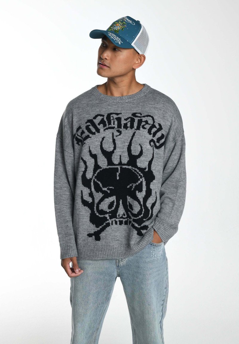 Grauer Pullover mit schwarzem Flammen- und Totenkopf-Muster, runder Ausschnitt und gerippten Säumen. Kombiniert mit hellblauen Jeans und einer blauen Mütze.