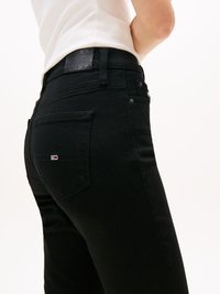 En person som bär svarta tajta jeans och en vit ärmlös topp, visad från midjan till mitten av låret mot en enfärgad bakgrund.