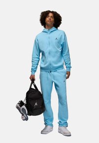 Błyszczący niebieski dres z kapturem na zamek i spodniami, z czarnym logo. Trzyma czarny sportowy plecak i białe trampki.