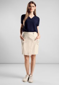 Marineblauwe v-hals blouse gecombineerd met een beige knielange rok met twee voorkeurszakken en een gebogen zoom. Het model draagt lichte sneakers.