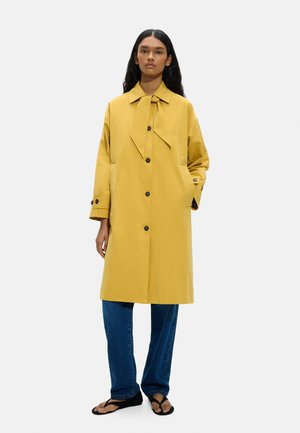 Kobieta ubrana w musztardowo-żółty płaszcz z guzikami i kołnierzem z kokardą, niebieskie dżinsy i czarne sandały, stojąca na jednolitym tle.