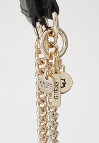 Cinturino in pelle nera con catene in tono oro, dotato di due targhette con incisioni "GUESS" e "G". Finitura lucida, design moderno.