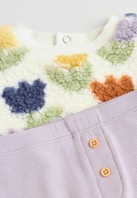 Fleece vršek s vícebarevným květinovým vzorem v odstínech žluté, zelené, oranžové a modré, spárovaný s levandulovým žebrovaným spodkem s dvěma dřevěnými knoflíky.