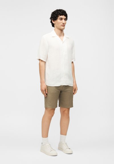 Άνδρας στέκεται φορώντας λευκό πουκάμισο με κοντά μανίκια και κουμπιά, καφέ σορτς μέχρι το γόνατο, λευκές κάλτσες και λευκά αθλητικά σε απλό φόντο.