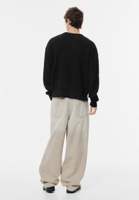 Maglione nero a coste, vestibilità ampia. Pantaloni denim beige chiaro a gamba larga con tasche posteriori. Scarpe nere. Sfondo minimalista.