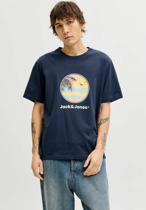 Młody mężczyzna noszący granatowy T-shirt Jack & Jones z grafiką zachodu słońca, liści palmowych i ptaków, w połączeniu z luźnymi niebieskimi jeansami.