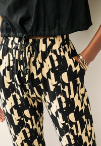 Person med højtaljede bukser i beige og sort abstrakt mønster og en sort cropped top, den ene hånd i lommen med guldfarvede armbånd.