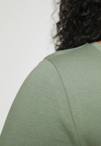 Vihreä lyhythihainen t-paita, jossa on ribbineulos ja sileä sauma olkapäällä, esittelee yksinkertaisia mallityylisiä yksityiskohtia.