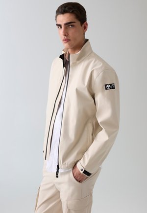 Joven de pie con las manos en los bolsillos, vistiendo una chaqueta beige con cremallera y pantalones a juego sobre un fondo claro y liso.