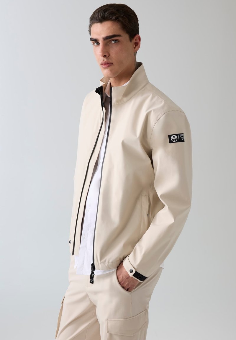 Jeune homme debout, mains dans les poches, portant une veste zippée beige et un pantalon assorti sur un fond clair uni.