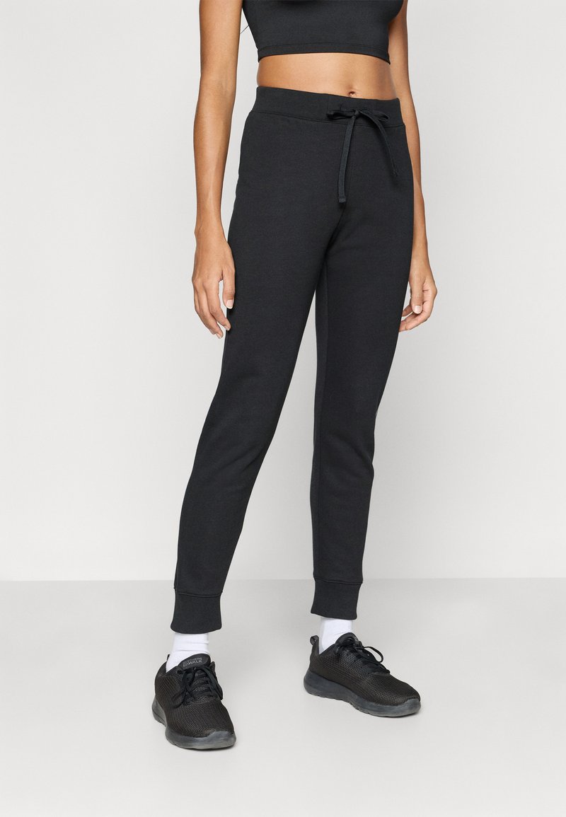 Μαύρο παντελόνι φόρμας με κορδόνι στη μέση, εφαρμοστά πόδια που στενεύουν και ρεβέρ στους αστραγάλους, φτιαγμένο από απαλό, υφασμένο ύφασμα. Φορεμένο με μαύρα αθλητικά παπούτσια.