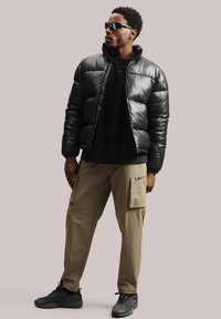 Melna pūkaina jaka, spīdīga materiāla, ar rāvējslēdzēju priekšpusē, garām piedurknēm. Melna krekla džemperis zem tās, tan krāsas cargo bikses ar sānu kabatām, melni sporta apavi.