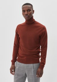 Vīrietis, kurš ģērbies koraļļu krāsā turtleneck džemperī un pelējos kareivēnē ar skaldām stāvu pretī vienkāršam gaiši pelēkam fona.