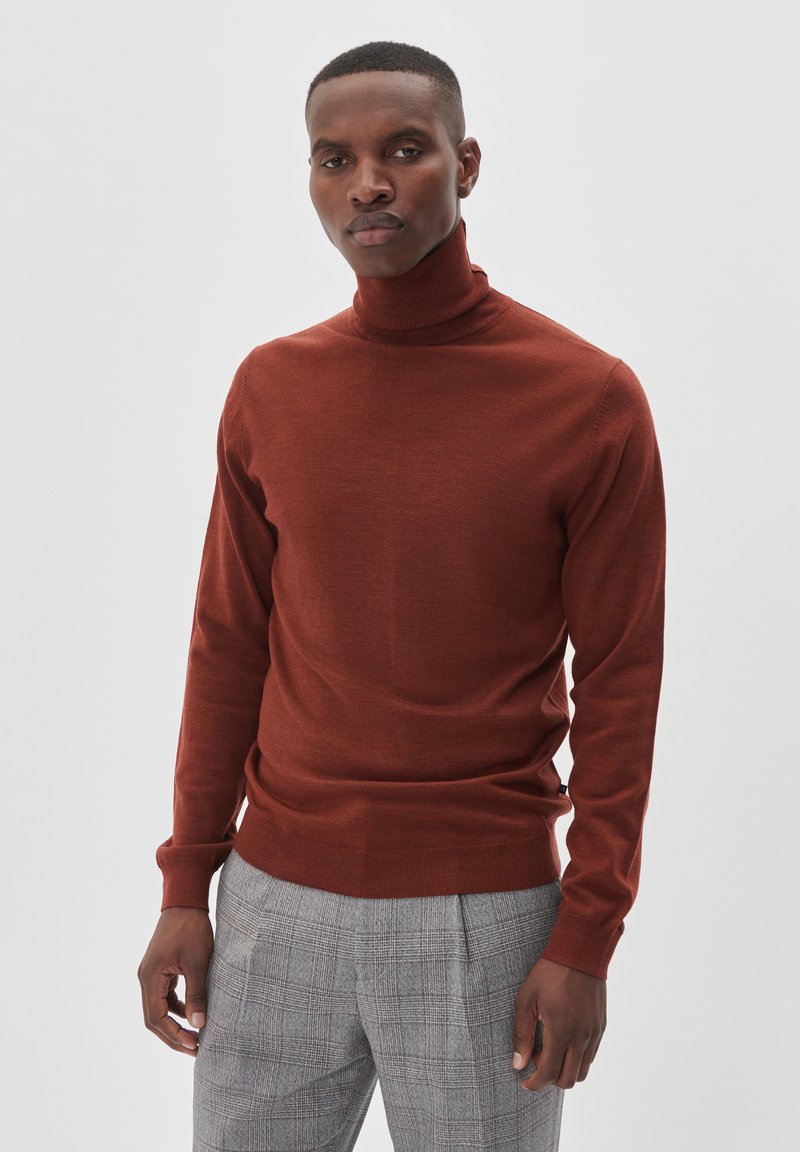 Vīrietis, kurš ģērbies koraļļu krāsā turtleneck džemperī un pelējos kareivēnē ar skaldām stāvu pretī vienkāršam gaiši pelēkam fona.