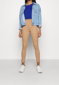 Ljusblå denimjacka över en blå skjorta, tillsammans med åtsittande tanbyxor och vita sneakers. Släta texturer och avslappnad design.