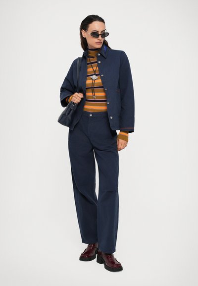 Γυναίκα που φοράει ναυτική ζακέτα και παντελόνι, ριγέ ζιβάγκο, μπορντώ ελαστικές πλατφόρμες, σκούρα γυαλιά ηλίου και κρατάει ναυτική τσάντα.