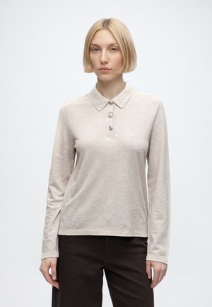 Jeune personne aux cheveux blonds courts portant un polo beige à manches longues et un pantalon foncé sur un fond clair uni.