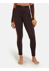 Vrouw in donkerbruine leggings met beige zijkanten en een bijpassend lange mouwen top, staande op blote voeten tegen een witte achtergrond.