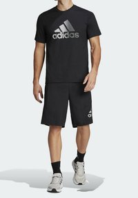 Musta urheiluasu, jossa on lyhythihainen t-paita, jossa on suuri heijastava Adidas-logo, sekä yhteensopivat shortsit, joiden kankaassa on tekstuuria ja rento istuvuus.