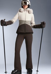 Skioutfit mit einer zweifarbigen Jacke in Creme und Grau mit Fellakzenten, kombiniert mit braunen Hosen und schwarzen Skischuhen, gehalten mit Skistöcken.