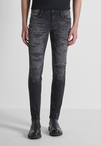 Jeans de denim negro y desgastado con un corte ajustado, que presentan patrones desvanecidos y un acabado suave y texturado. Bolsillos frontales y detalles metálicos.