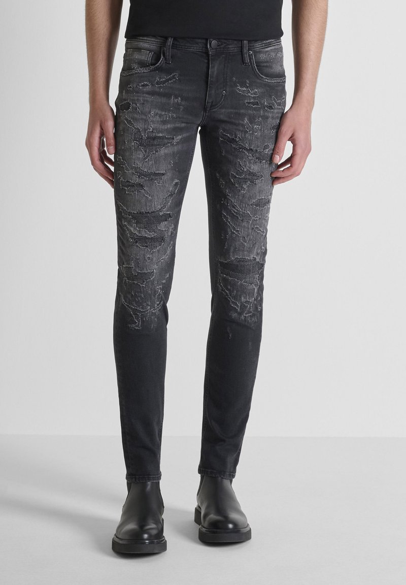Jeans de denim negro y desgastado con un corte ajustado, que presentan patrones desvanecidos y un acabado suave y texturado. Bolsillos frontales y detalles metálicos.