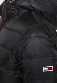 Chaqueta acolchada negra con una textura brillante y acolchada, con cierre de cremallera y un parche de logo prominente en rojo, blanco y azul en la manga.