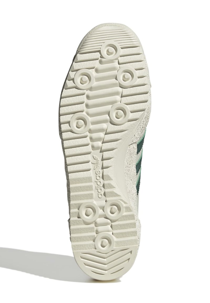 Semelle blanche avec une surface texturée, présentant des grips circulaires et un logo discret. Trois bandes vertes sur le côté et un matériau supérieur gris.
