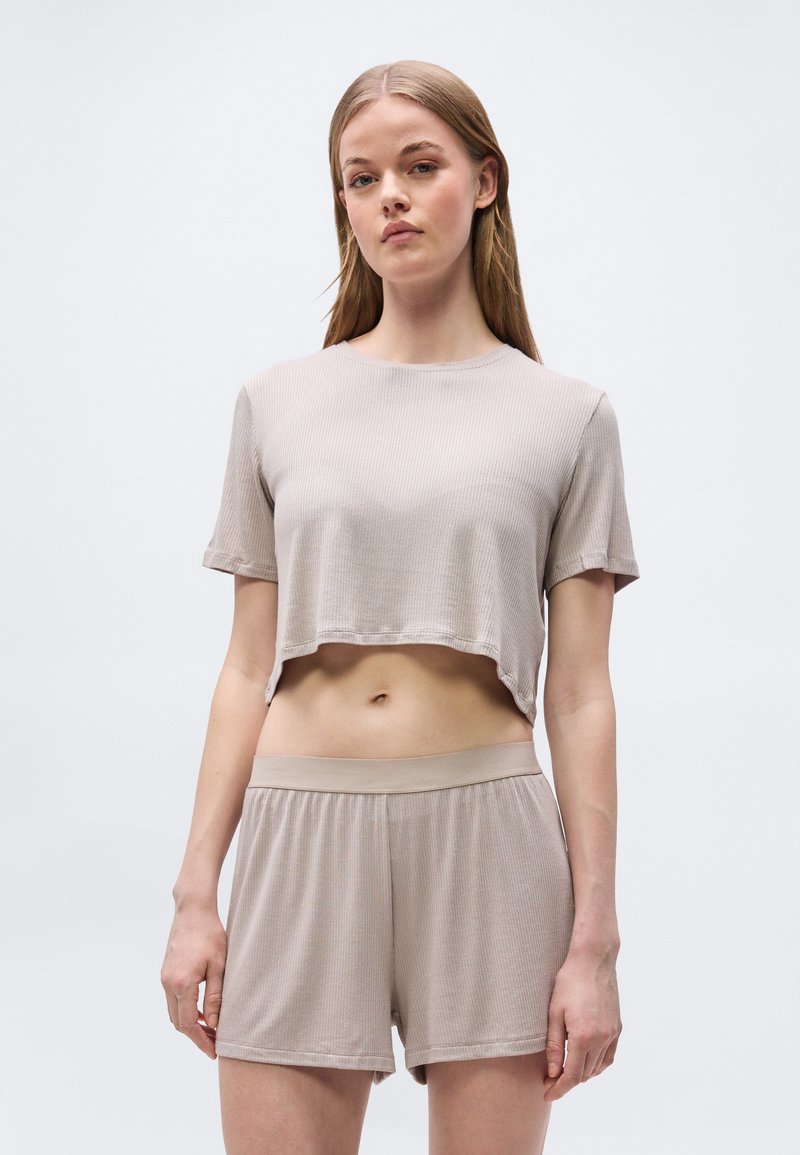 Νεαρή γυναίκα όρθια, φοράει μπεζ ριμπ κοντομάνικο τοπ και κοντά σορτς με ελαστική μέση, ουδέτερο φόντο.