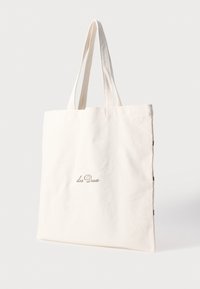 Καμβάς τσάντα ώμου σε κρεμ χρώμα με κείμενο "Les Deux" σε ανοιχτό καφέ καλλιγραφία. Δύο ανθεκτικές λαβές και ένα απλό, χωρίς στολίδια σχέδιο.