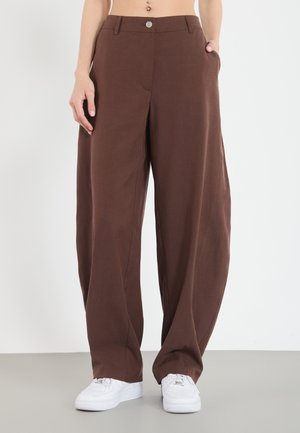 Brown, loose-fitting trousers with a smooth texture, featuring side pockets and a front button closure, paired with white sneakers.