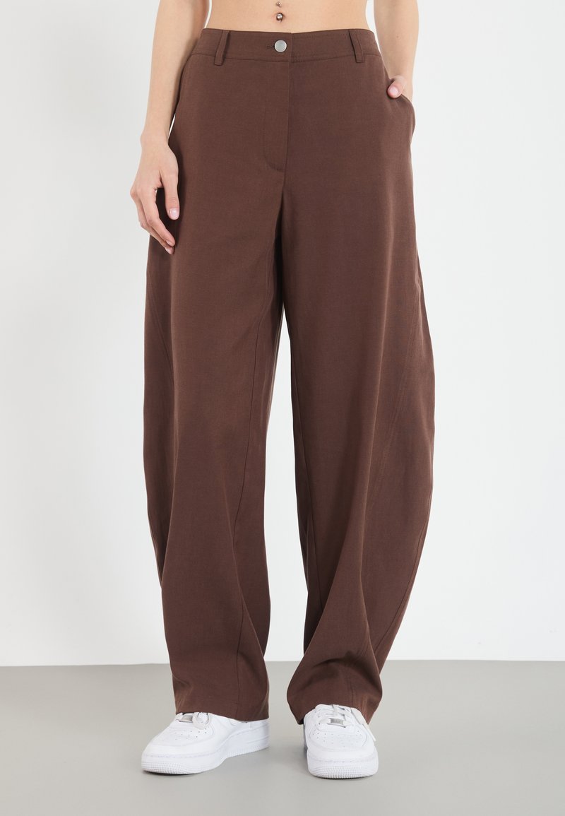 Pantalons marron à coupe ample avec une texture lisse, dotés de poches latérales et d'un bouton de fermeture à l'avant, associés à des baskets blanches.