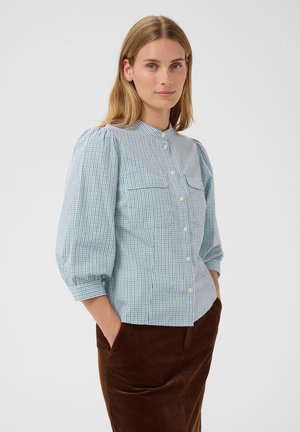 Frau mit hellbraunem Haar, die eine hellblaue karierte Bluse mit Puffärmeln und braune Cordhose trägt, mit Händen in den Taschen.