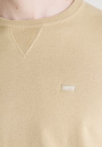 Close-up van een beige crewneck sweatshirt met ribbel V-stiksel detail en een klein wit Levi's logo op de borst.